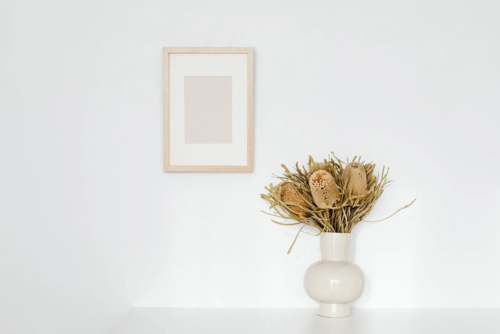 Simple white wall interior with framed picture and ceramic vase holding dried flowers.